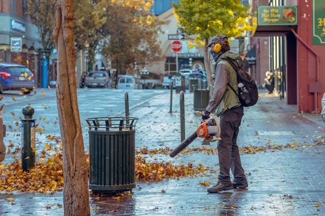 A worker using a blower to remove leaves from the sidewalk